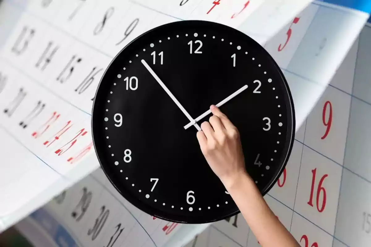 Una mano ajusta las manecillas de un reloj negro sobre un fondo de calendario.