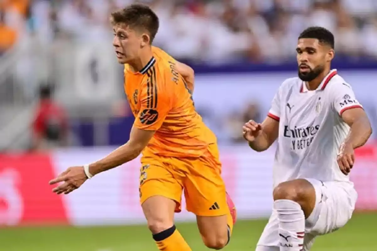 Dos jugadores de fútbol en acción durante un partido, uno con uniforme naranja y el otro con uniforme blanco.