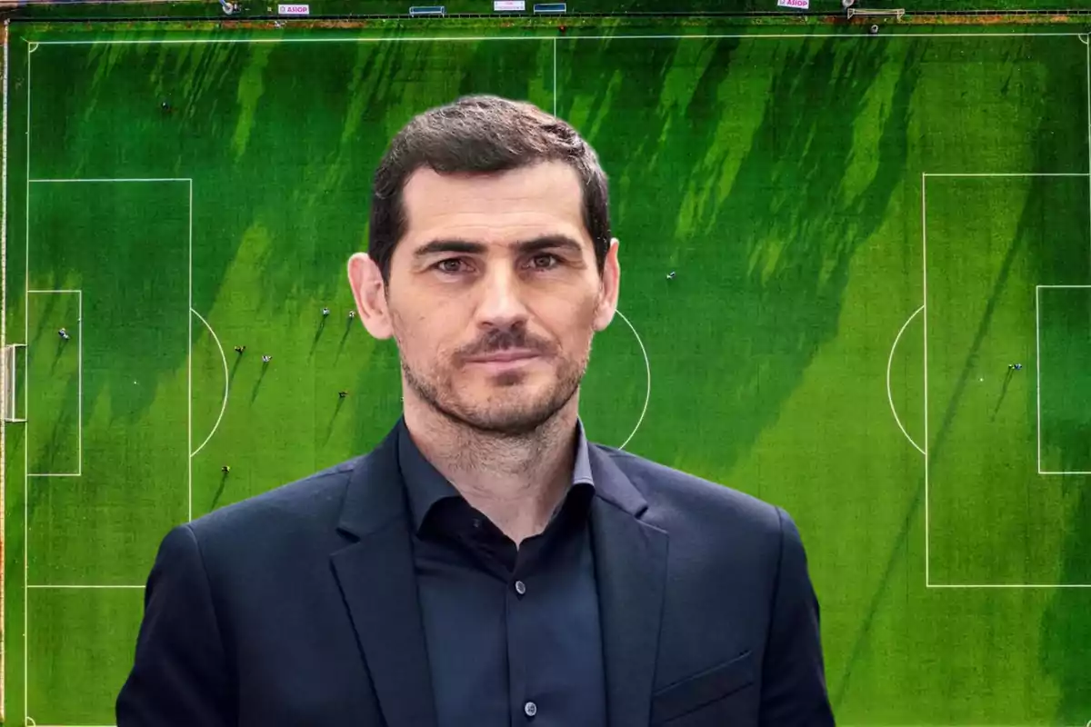 Hombre de cabello corto y barba con traje oscuro posando frente a un campo de fútbol visto desde arriba