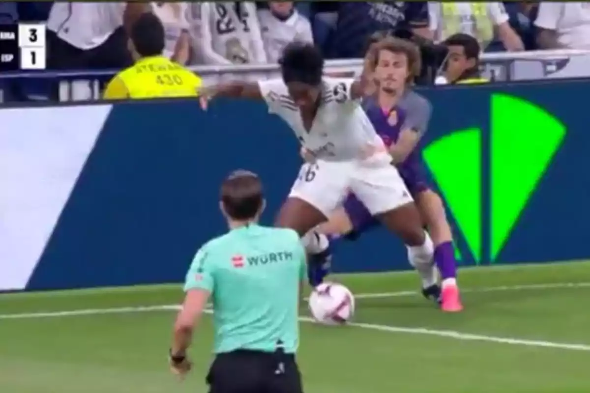 Un jugador de fútbol con uniforme blanco está siendo marcado por un oponente con uniforme morado mientras un árbitro observa la jugada.