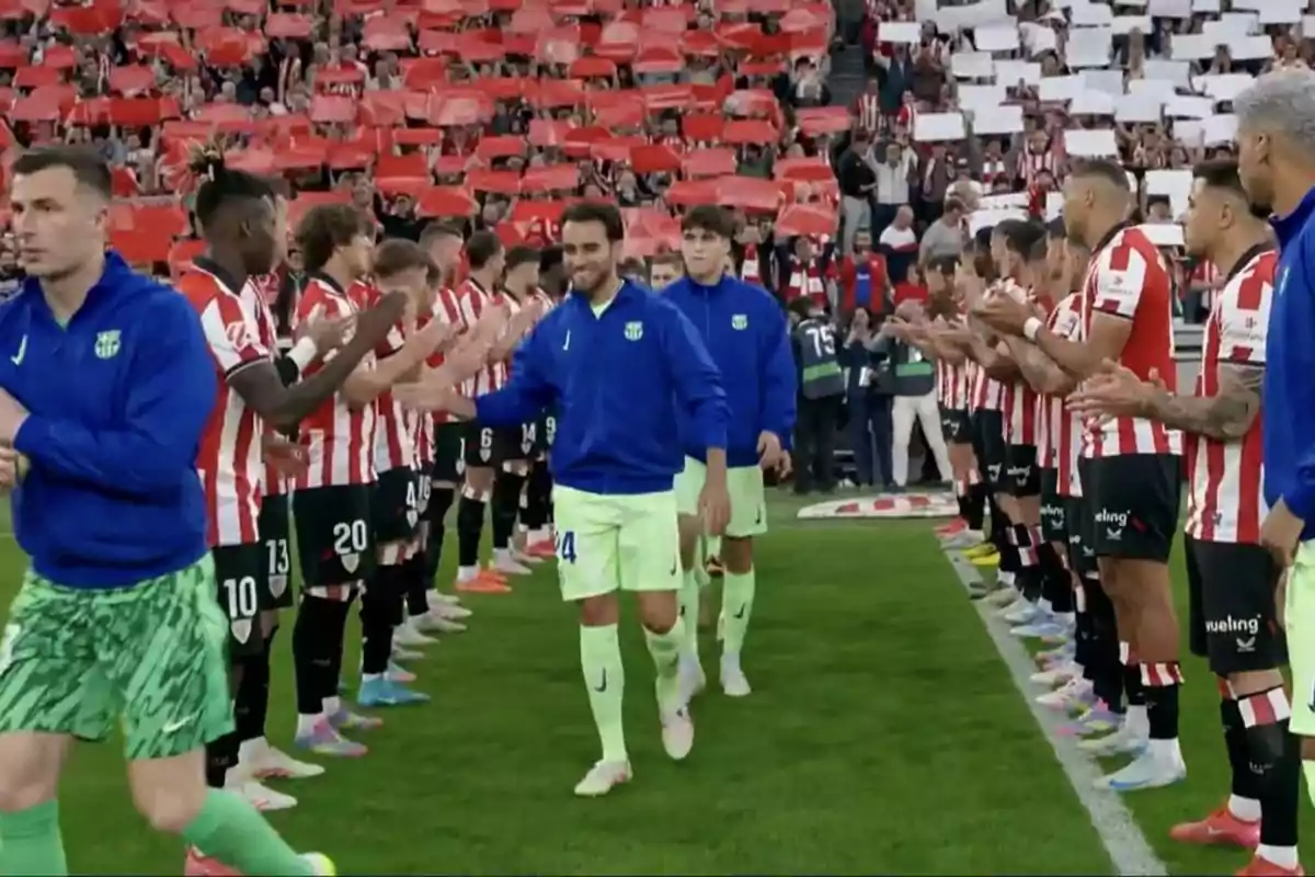 Jugadores de un equipo de fútbol forman un pasillo y aplauden mientras los jugadores del equipo rival caminan entre ellos en un estadio lleno de aficionados que sostienen cartulinas rojas y blancas Jugadores de un equipo de fútbol forman un pasillo y aplauden mientras los jugadores del equipo rival caminan entre ellos en un estadio lleno de aficionados que sostienen cartulinas rojas y blancas