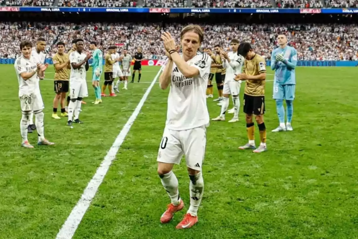 Un jugador de fútbol aplaude mientras camina por el campo, rodeado de compañeros y rivales que también aplauden, con un estadio lleno de espectadores al fondo. Un jugador de fútbol aplaude mientras camina por el campo, rodeado de compañeros y rivales que también aplauden, con un estadio lleno de espectadores al fondo.