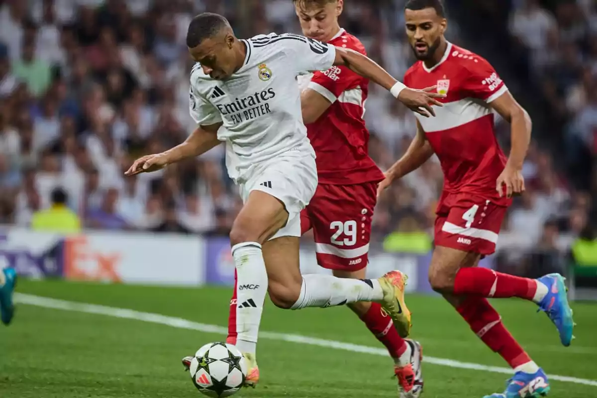 Un jugador de fútbol del Real Madrid en acción durante un partido, siendo marcado por dos jugadores del equipo contrario vestidos de rojo.