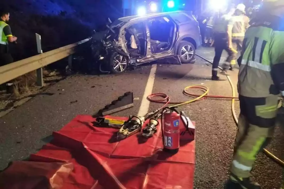 Accidente de tráfico en carretera con un coche gravemente dañado, presencia de bomberos, herramientas de rescate y un extintor sobre una manta roja Accidente de tráfico en carretera con un coche gravemente dañado, presencia de bomberos, herramientas de rescate y un extintor sobre una manta roja