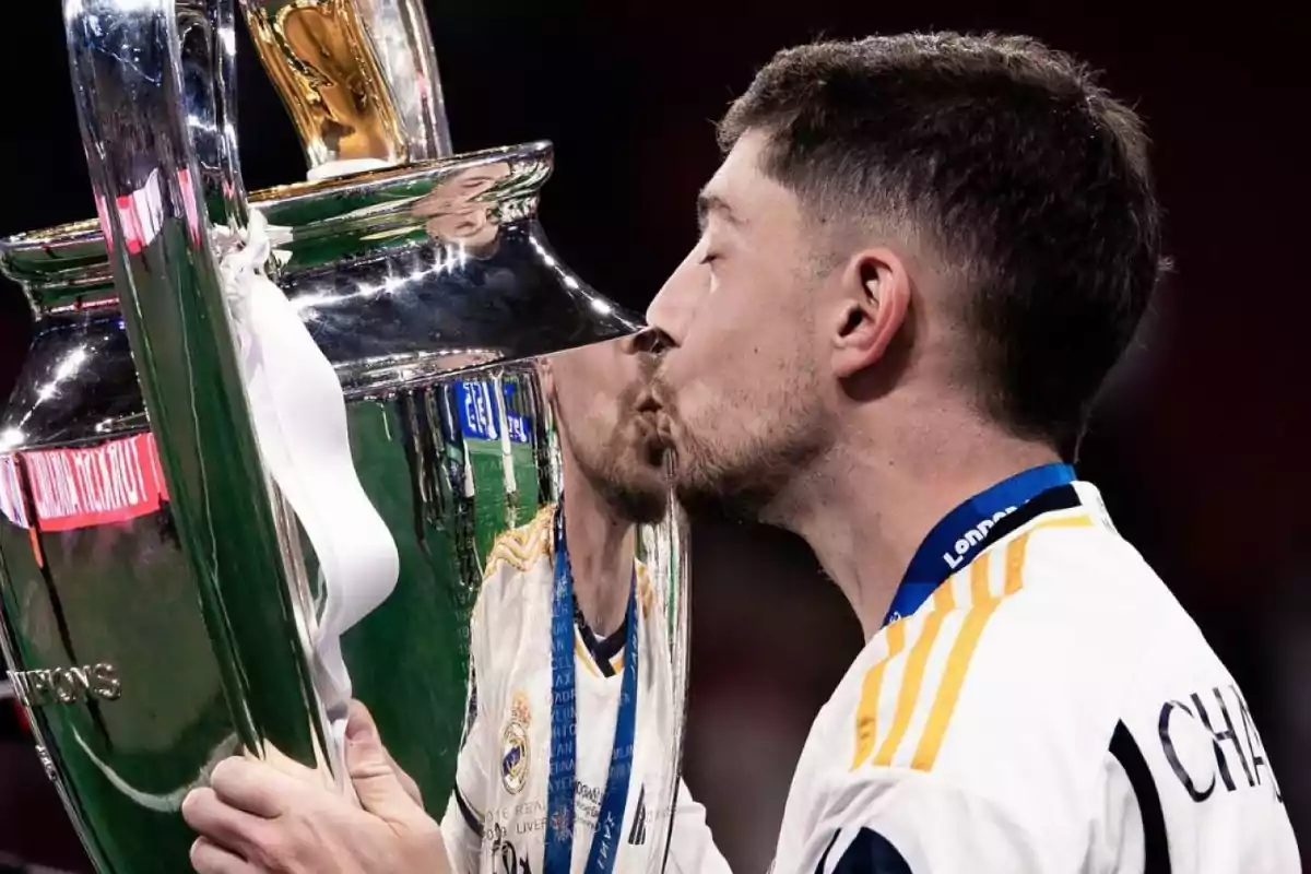 Un jugador de fútbol besa un trofeo grande y plateado mientras lleva una medalla alrededor del cuello.