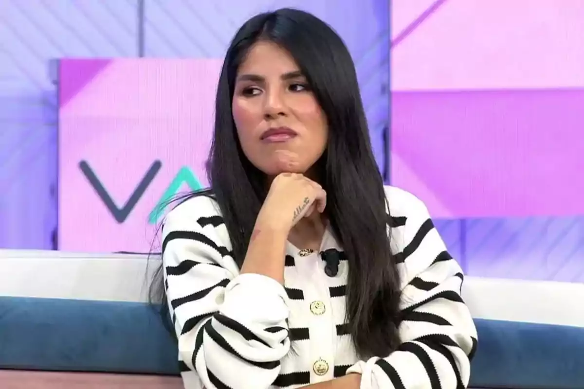 Una mujer con cabello largo y oscuro está sentada en un set de televisión con fondo colorido, lleva un suéter de rayas blancas y negras y apoya su barbilla en su mano.