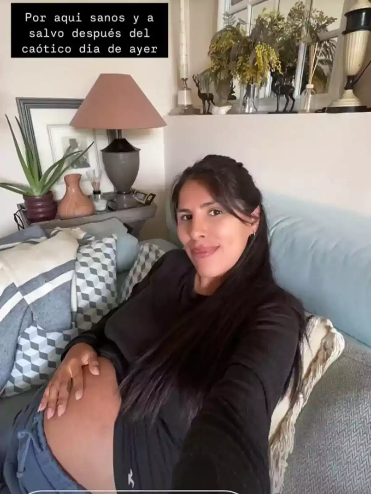 Una mujer embarazada está sentada en un sofá, sonriendo y tocando su vientre, con una decoración hogareña de fondo que incluye plantas y una lámpara. Una mujer embarazada está sentada en un sofá, sonriendo y tocando su vientre, con una decoración hogareña de fondo que incluye plantas y una lámpara.