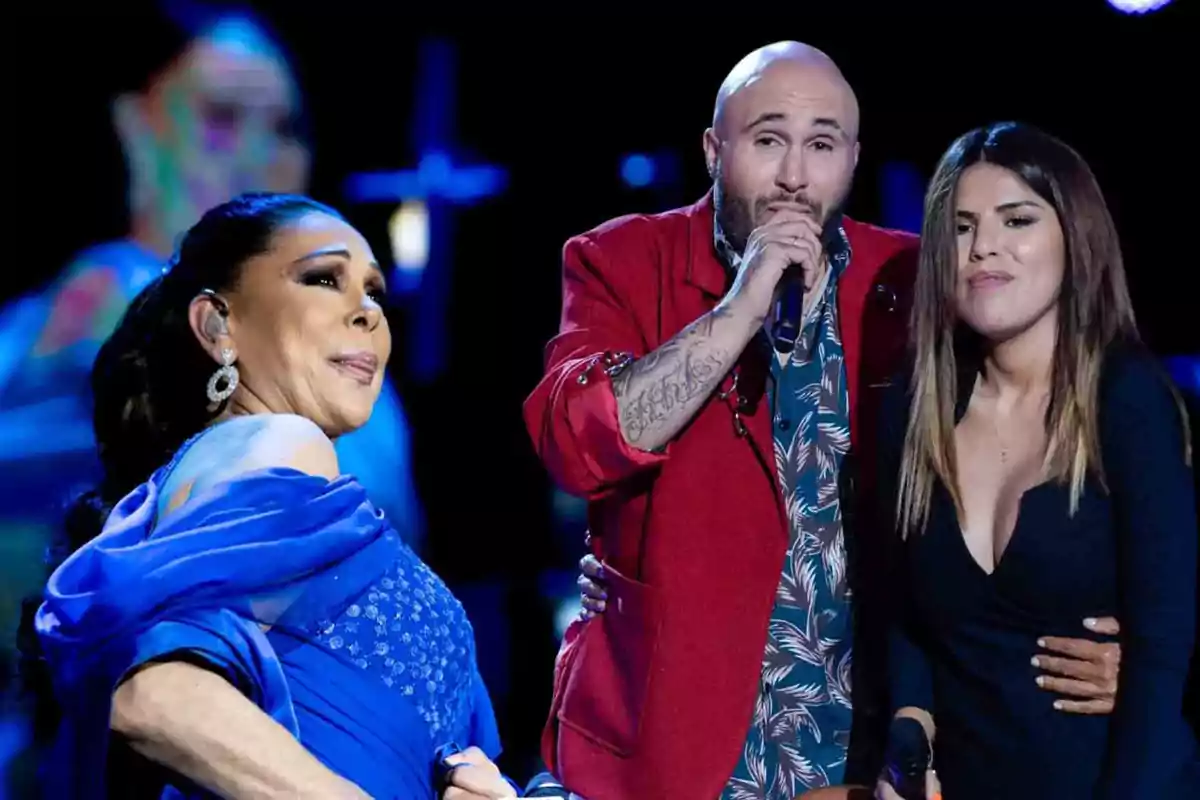 Tres personas en un escenario durante una presentación musical, una mujer con vestido azul, un hombre con chaqueta roja y una mujer con vestido negro.