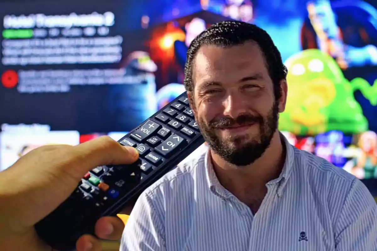 Una persona sonriendo frente a una pantalla de televisión con una mano sosteniendo un control remoto.