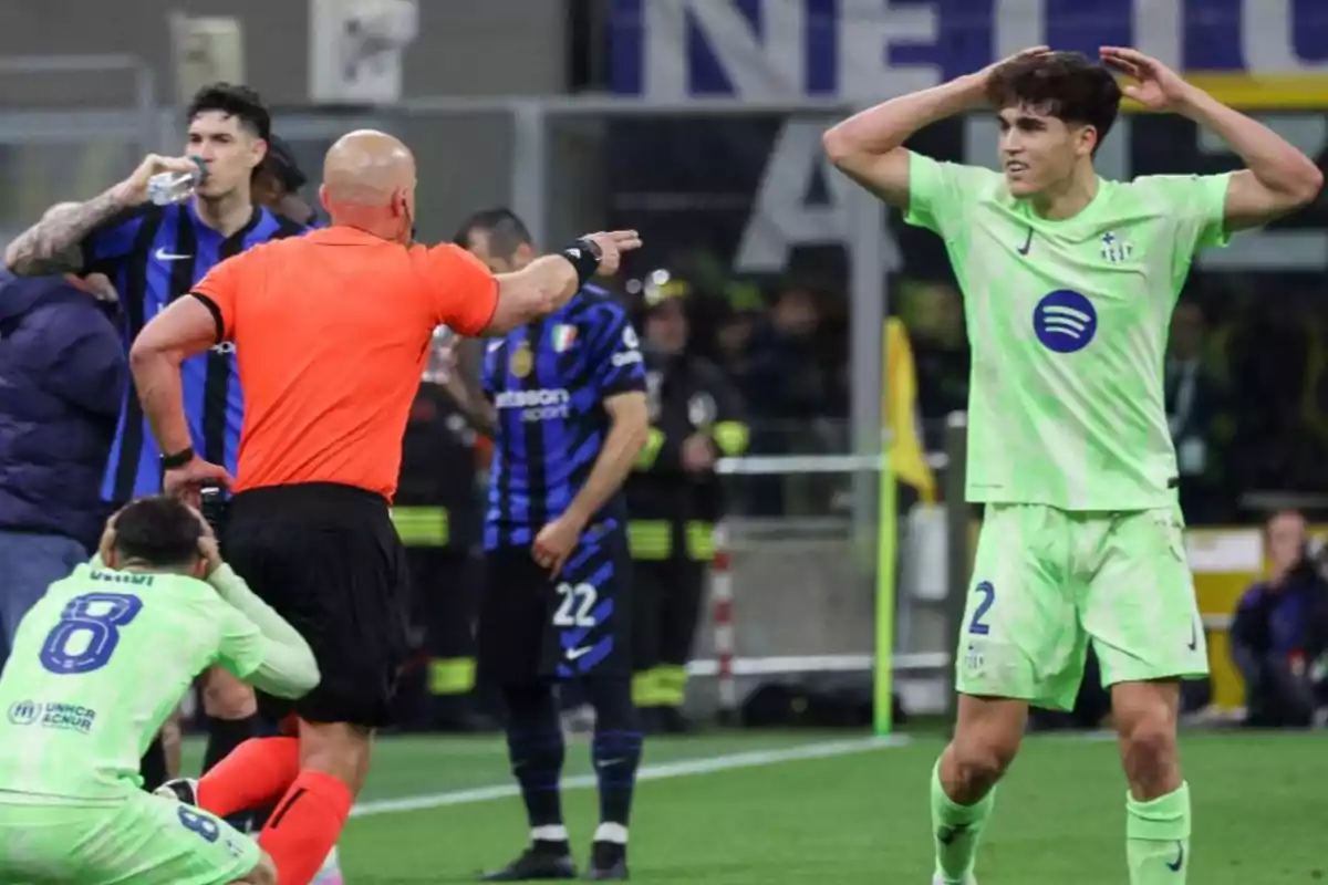 Un árbitro señala una decisión en el campo mientras un jugador de verde se lleva las manos a la cabeza y otro está arrodillado, con jugadores del equipo contrario observando la escena. Un árbitro señala una decisión en el campo mientras un jugador de verde se lleva las manos a la cabeza y otro está arrodillado, con jugadores del equipo contrario observando la escena.