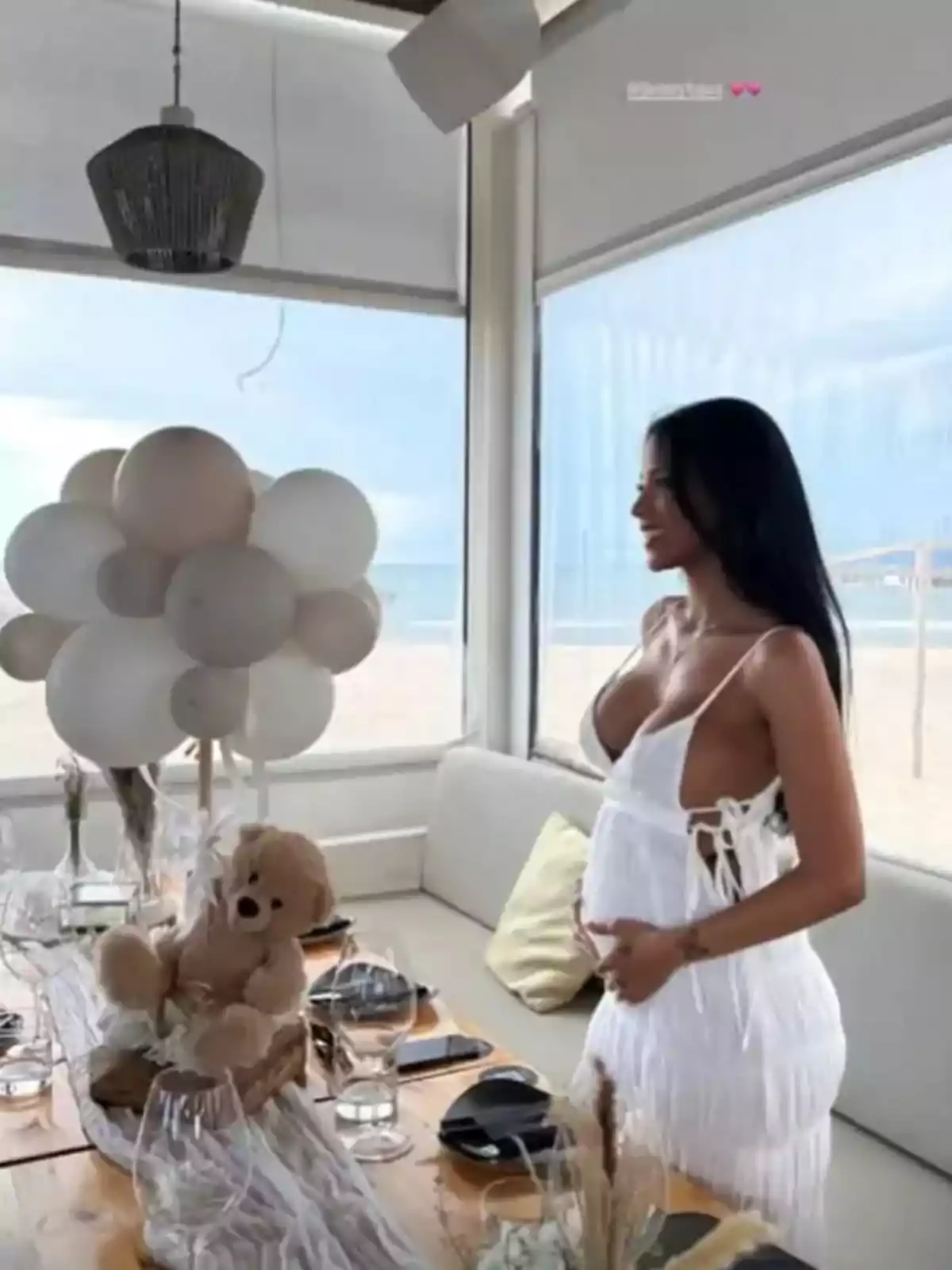 Una mujer embarazada vestida de blanco está de pie junto a una mesa decorada con globos blancos y un oso de peluche en un ambiente con vista a la playa. Una mujer embarazada vestida de blanco está de pie junto a una mesa decorada con globos blancos y un oso de peluche en un ambiente con vista a la playa.