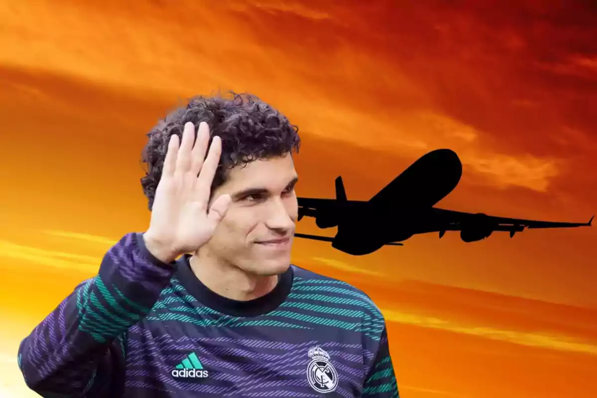 Un hombre con una camiseta de fútbol del Real Madrid levanta la mano en señal de saludo mientras un avión vuela en el cielo al atardecer.