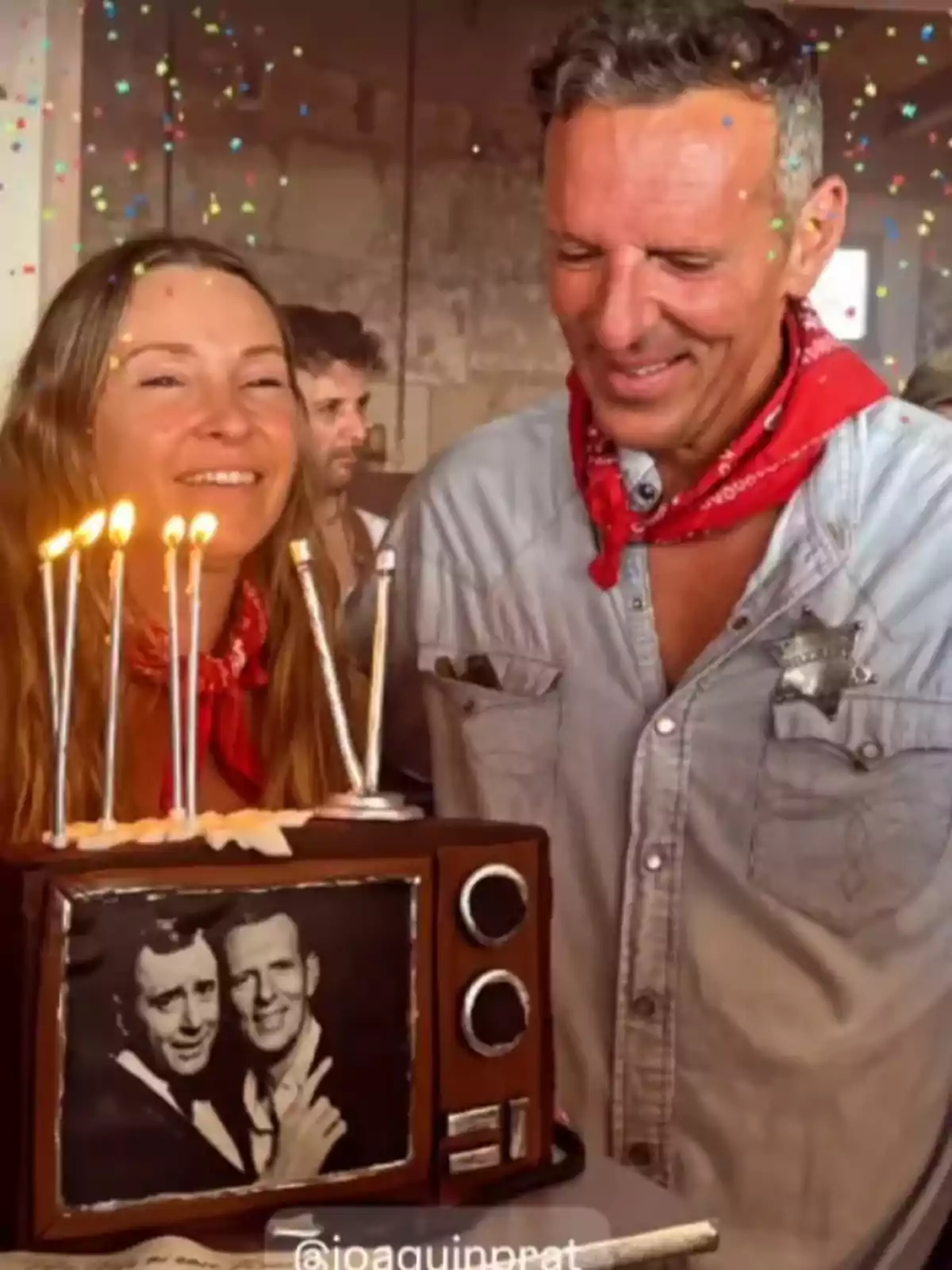 Una mujer y un hombre sonríen mientras sostienen un pastel con forma de televisor antiguo, decorado con velas encendidas y una imagen en blanco y negro en la pantalla, mientras confeti cae alrededor. Una mujer y un hombre sonríen mientras sostienen un pastel con forma de televisor antiguo, decorado con velas encendidas y una imagen en blanco y negro en la pantalla, mientras confeti cae alrededor.