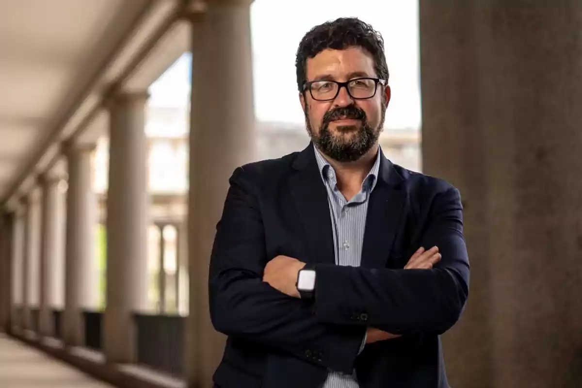 Hombre con barba y gafas, vestido con traje oscuro y camisa clara, posando con los brazos cruzados en un pasillo con columnas. Hombre con barba y gafas, vestido con traje oscuro y camisa clara, posando con los brazos cruzados en un pasillo con columnas.