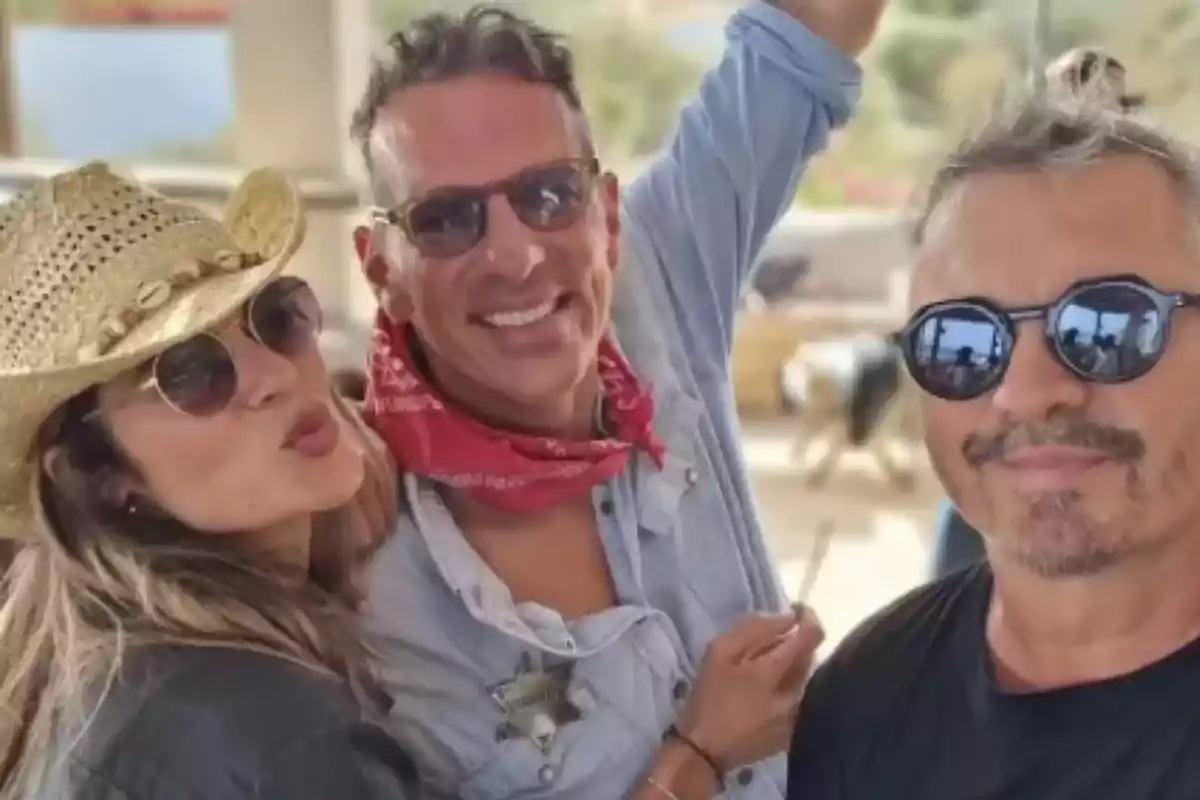 Tres personas posan juntas sonriendo; una mujer lleva un sombrero de vaquero y gafas de sol, el hombre en el centro lleva una camisa de mezclilla y un pañuelo rojo, y el hombre a la derecha lleva gafas de sol y una camiseta negra. Tres personas posan juntas sonriendo; una mujer lleva un sombrero de vaquero y gafas de sol, el hombre en el centro lleva una camisa de mezclilla y un pañuelo rojo, y el hombre a la derecha lleva gafas de sol y una camiseta negra.