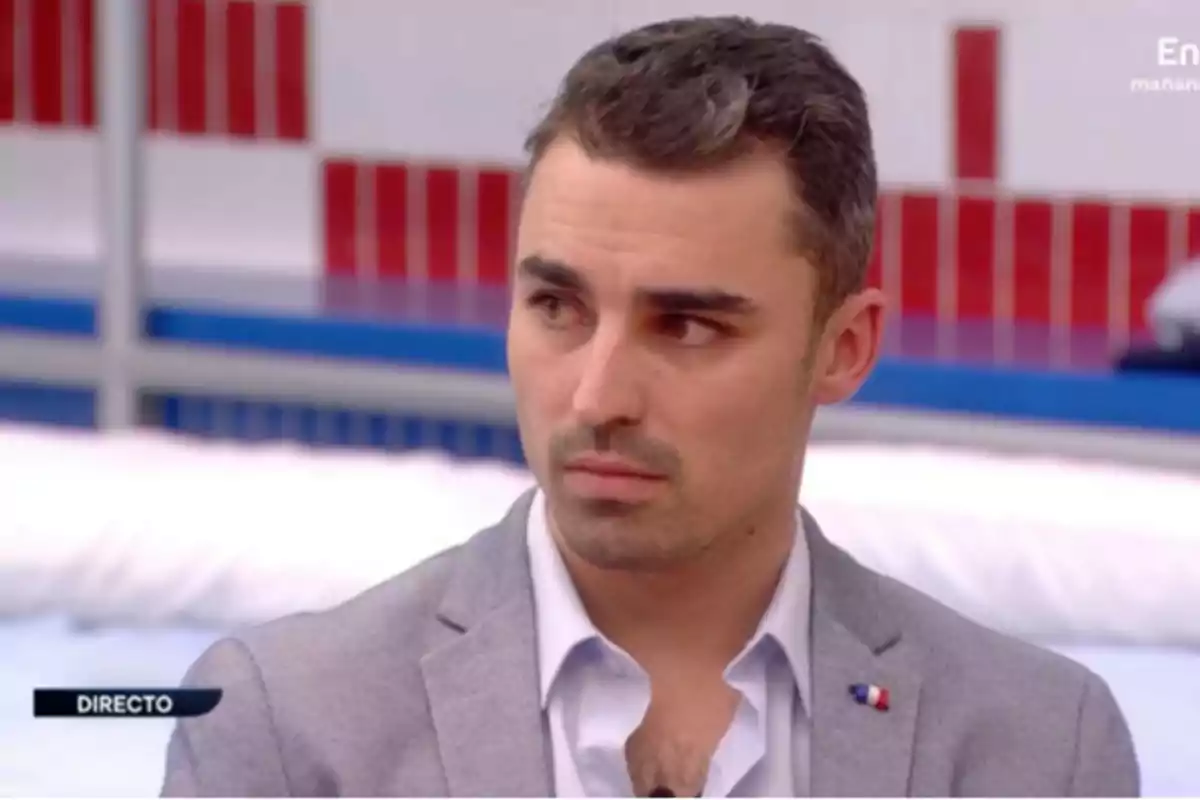 Un hombre con cabello corto y barba incipiente, vestido con una camisa azul clara y un saco gris, aparece en una transmisión en vivo con un fondo de colores rojo, blanco y azul. Un hombre con cabello corto y barba incipiente, vestido con una camisa azul clara y un saco gris, aparece en una transmisión en vivo con un fondo de colores rojo, blanco y azul.