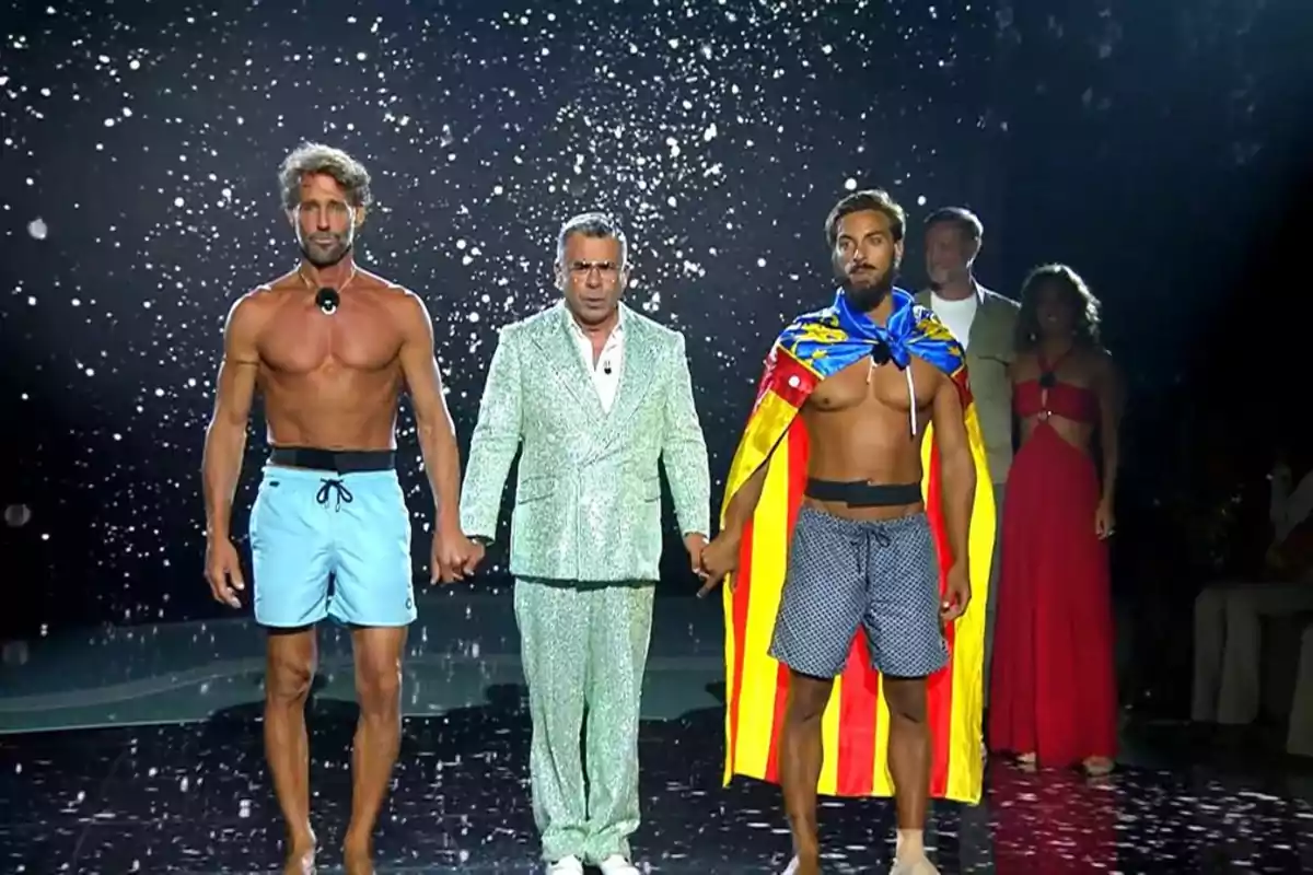 Tres hombres de pie en un escenario, dos de ellos en bañador y uno con traje brillante, mientras uno lleva una bandera sobre los hombros y de fondo caen destellos blancos Tres hombres de pie en un escenario, dos de ellos en bañador y uno con traje brillante, mientras uno lleva una bandera sobre los hombros y de fondo caen destellos blancos