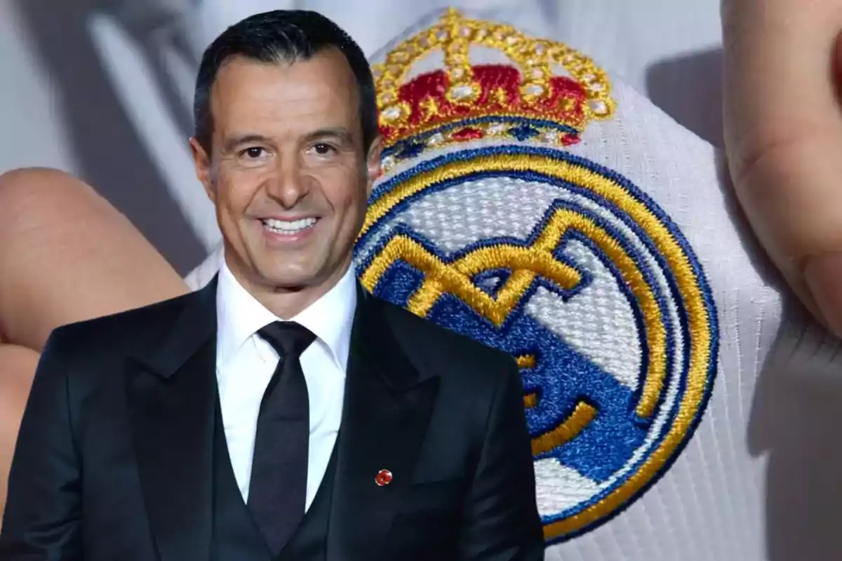 Un hombre con traje negro y corbata negra sonriendo frente a un escudo del Real Madrid.