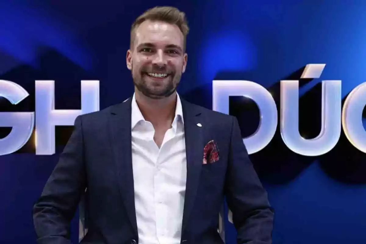 Un hombre sonriente con traje oscuro y camisa blanca frente a un fondo azul con letras grandes.