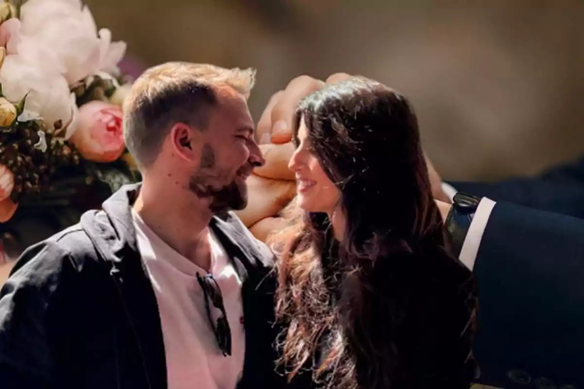 Una pareja sonriente se mira con afecto mientras un ramo de flores y manos entrelazadas se ven de fondo.