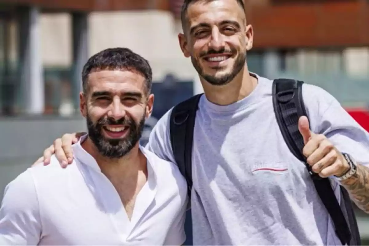 Dos hombres sonrientes posan juntos al aire libre, uno de ellos lleva una camiseta blanca y el otro una camiseta gris con una mochila.