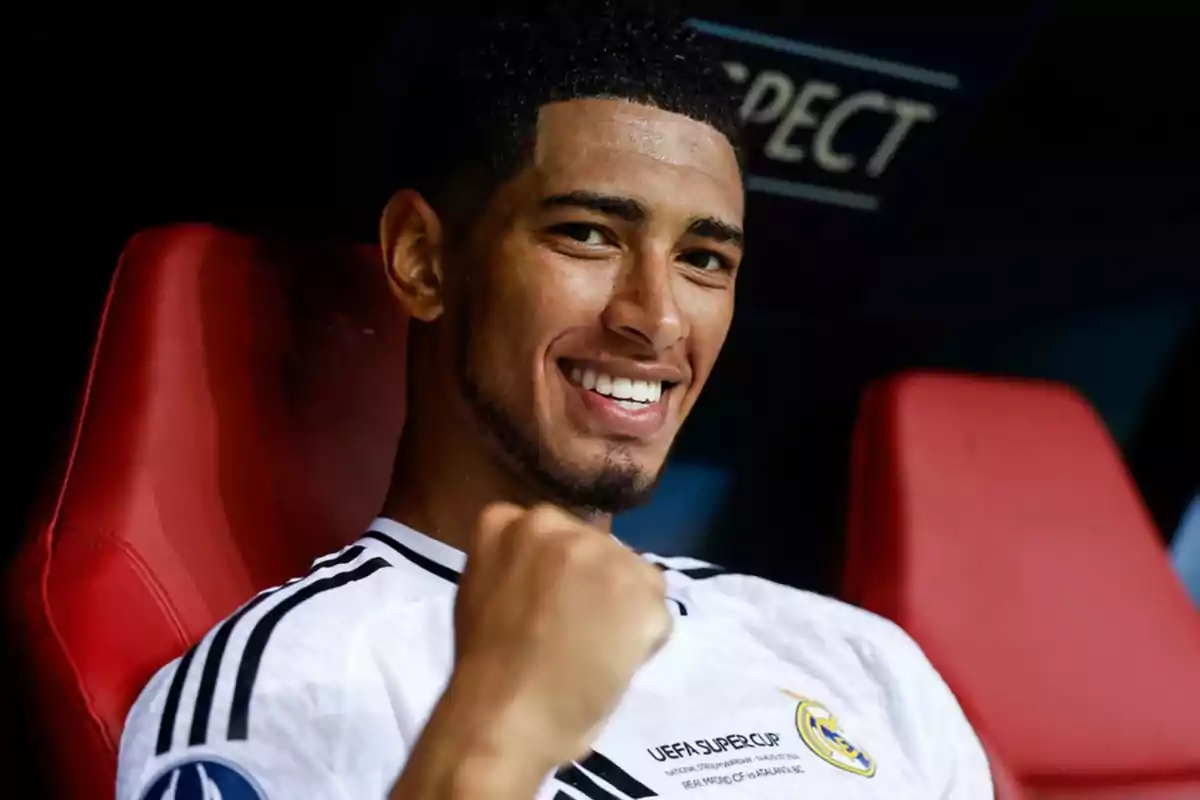 Un jugador de fútbol sonríe y levanta el puño mientras está sentado en un asiento rojo, vistiendo una camiseta blanca con el logo de la UEFA Super Cup.