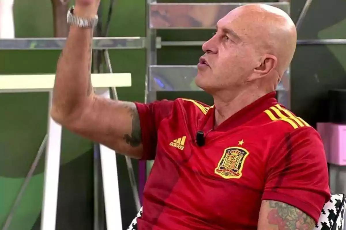 Un hombre calvo con una camiseta roja de la selección española de fútbol gesticulando con la mano en un estudio de televisión.