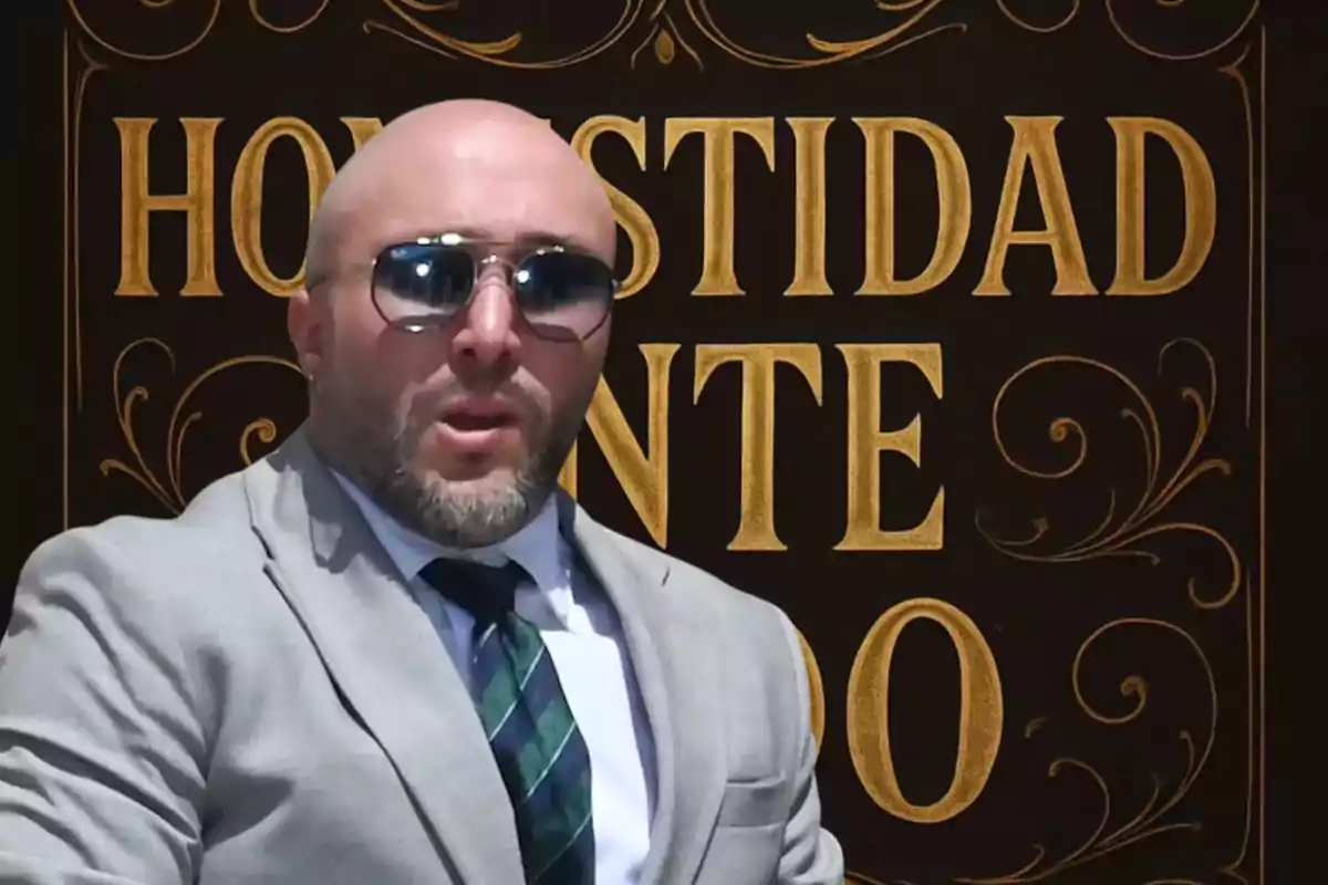 Hombre calvo con barba y gafas de sol vistiendo traje claro y corbata, posando frente a un fondo oscuro con letras doradas que dicen "Honestidad" y "Oro"