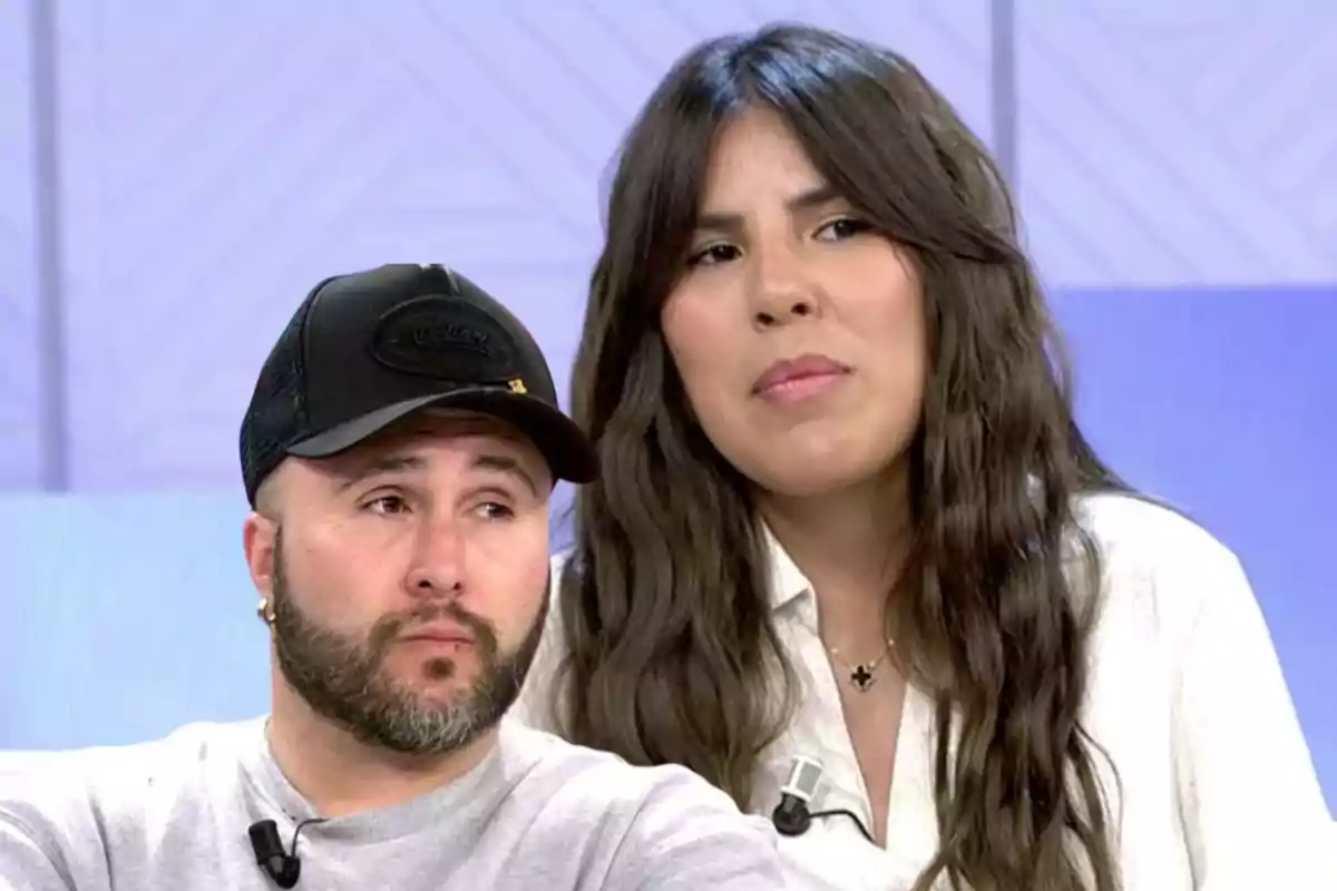 Dos personas en un programa de televisión, una mujer con cabello largo y una camisa blanca y un hombre con barba y gorra negra.