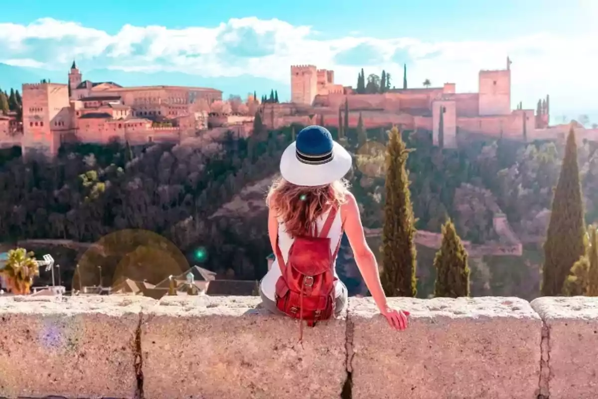 Persona con sombrero y mochila roja sentada en un muro contemplando un castillo histórico rodeado de árboles y montañas bajo un cielo azul.