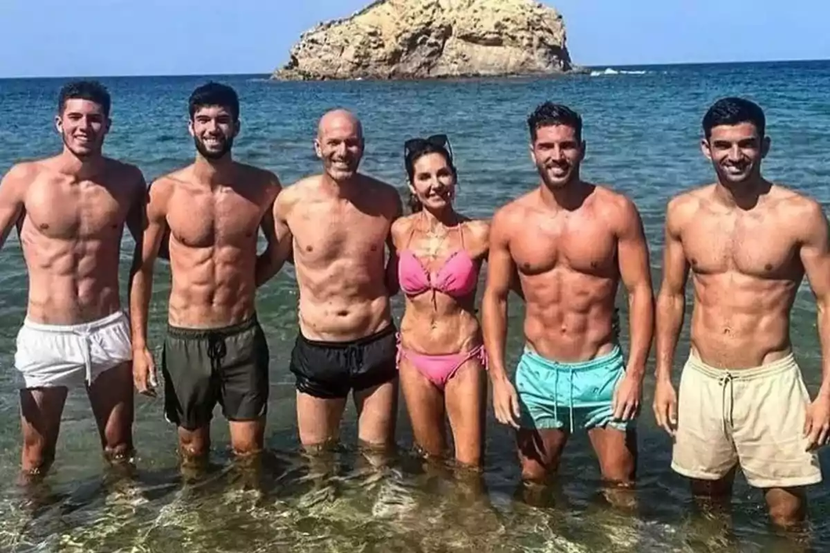 Un grupo de personas en traje de baño posando en la playa con una roca grande en el fondo.