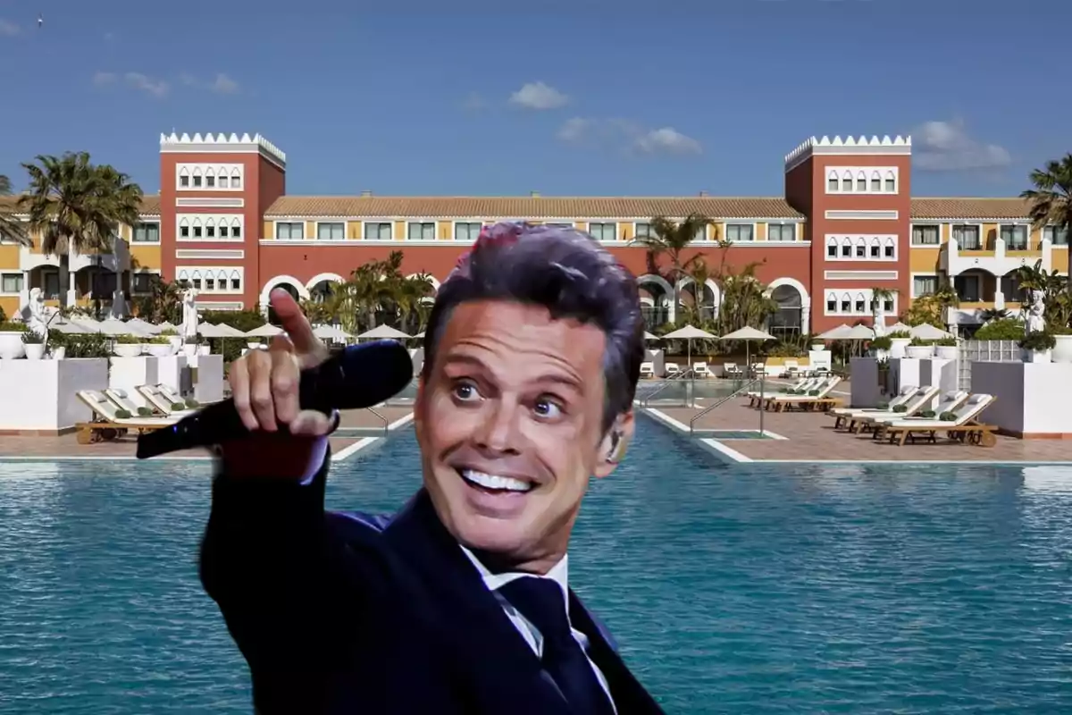 Un hombre sonriente con un micrófono en la mano frente a una piscina y un edificio de estilo mediterráneo.