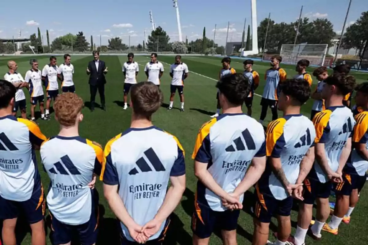 Un grupo de jugadores de fútbol escucha a un hombre que les habla en un campo de entrenamiento.