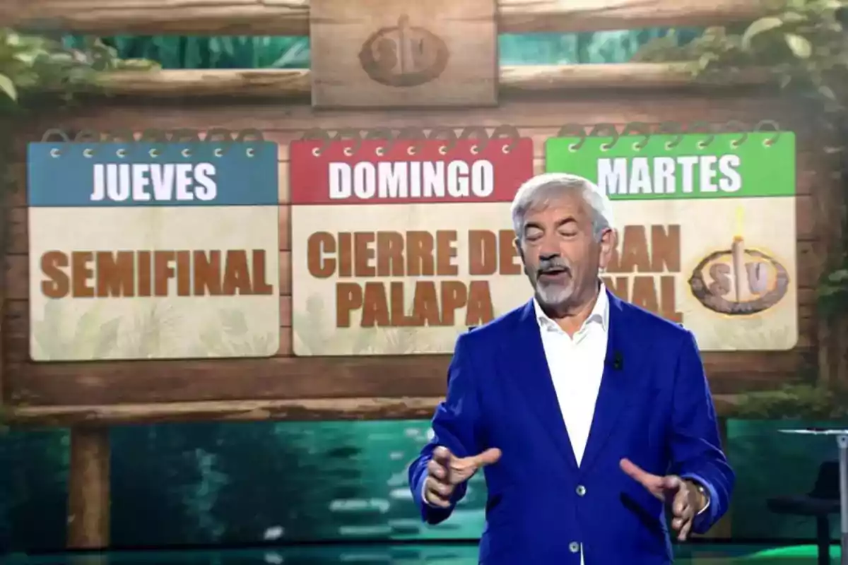 Un hombre de cabello canoso y barba, vestido con saco azul, está hablando frente a un cartel que anuncia los eventos de un programa de televisión con las palabras jueves semifinal, domingo cierre de palapa y martes gran final. Un hombre de cabello canoso y barba, vestido con saco azul, está hablando frente a un cartel que anuncia los eventos de un programa de televisión con las palabras jueves semifinal, domingo cierre de palapa y martes gran final.
