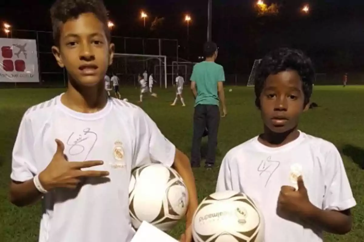 Dos niños con camisetas del Real Madrid sosteniendo balones de fútbol en un campo de entrenamiento nocturno.