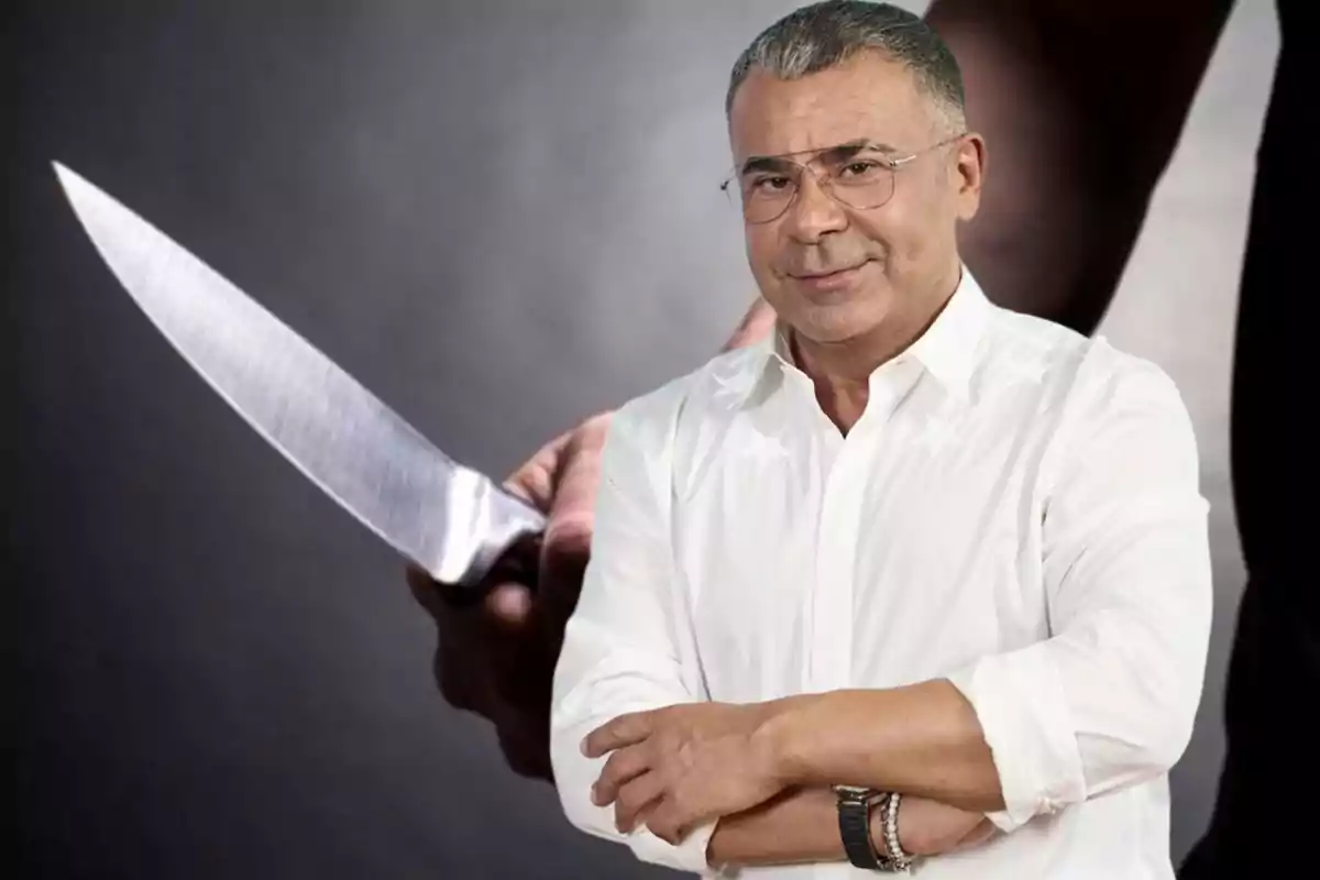 Un hombre con camisa blanca y gafas está de pie con los brazos cruzados, mientras que en el fondo se ve una mano sosteniendo un cuchillo grande.