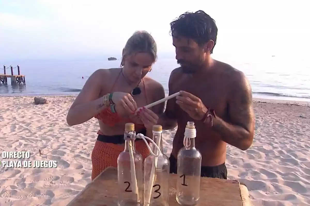 Dos personas en la playa observan un objeto mientras están junto a una mesa con botellas numeradas. Dos personas en la playa observan un objeto mientras están junto a una mesa con botellas numeradas.