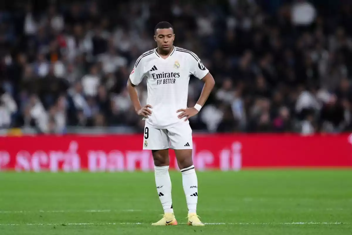 Un jugador de fútbol con uniforme blanco en el campo, con las manos en la cintura y una expresión pensativa.