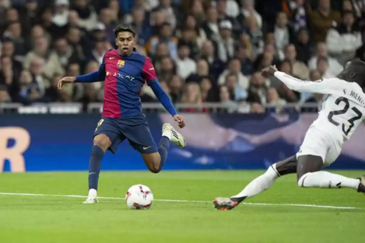 Dos jugadores de fútbol compiten por el balón en un partido, uno con uniforme azul y rojo y el otro con uniforme blanco, en un estadio lleno de espectadores.