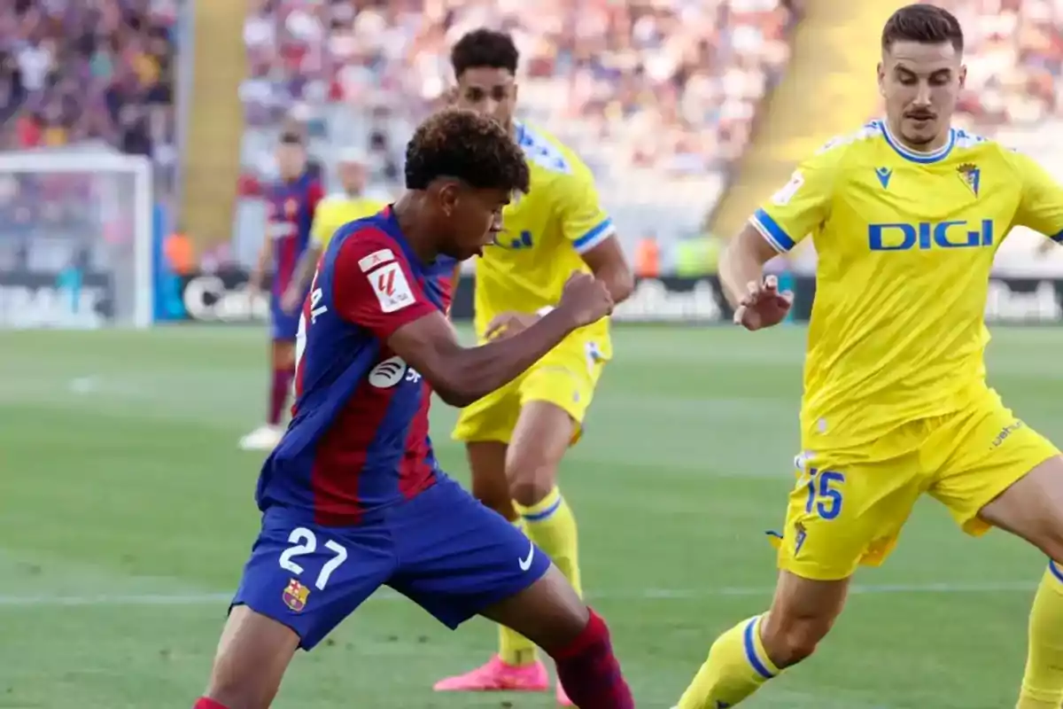 Jugadores de fútbol en acción durante un partido, uno con uniforme azul y rojo y los otros con uniforme amarillo.