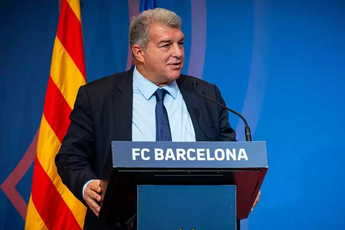 Hombre de traje y corbata hablando en un podio con el logo del FC Barcelona y una bandera catalana en el fondo.