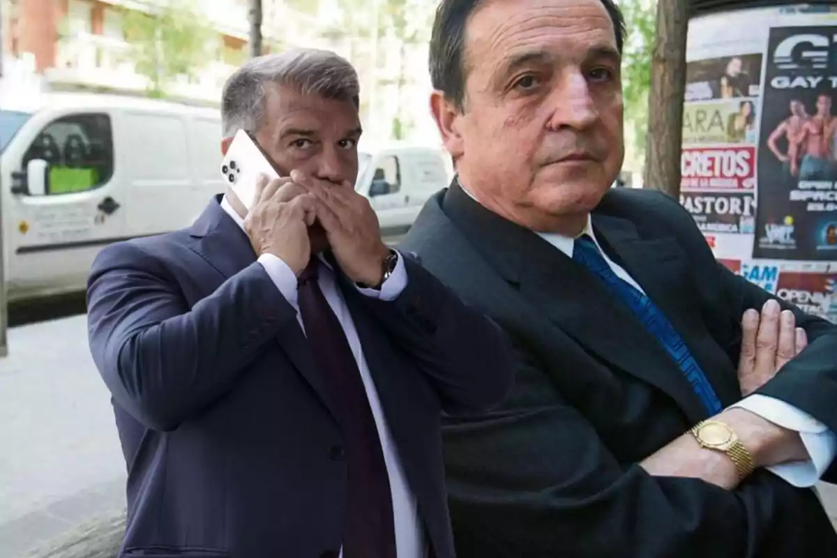 Dos hombres con traje posan en la calle, uno de ellos habla por teléfono móvil y el otro está de brazos cruzados frente a carteles publicitarios.