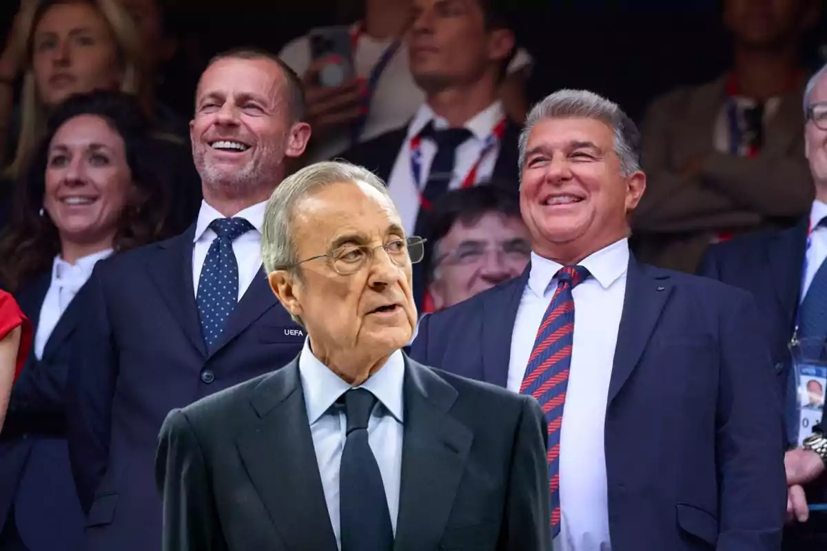 Un hombre mayor con gafas y traje oscuro en primer plano mientras dos hombres sonrientes con traje y corbata están al fondo rodeados de más personas en un evento formal