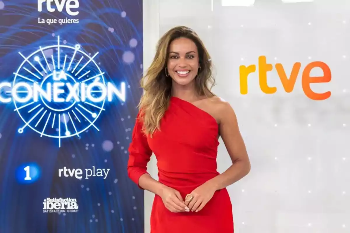 Mujer con vestido rojo de un solo hombro sonriendo frente a un fondo con logotipos de RTVE y La Conexión Mujer con vestido rojo de un solo hombro sonriendo frente a un fondo con logotipos de RTVE y La Conexión