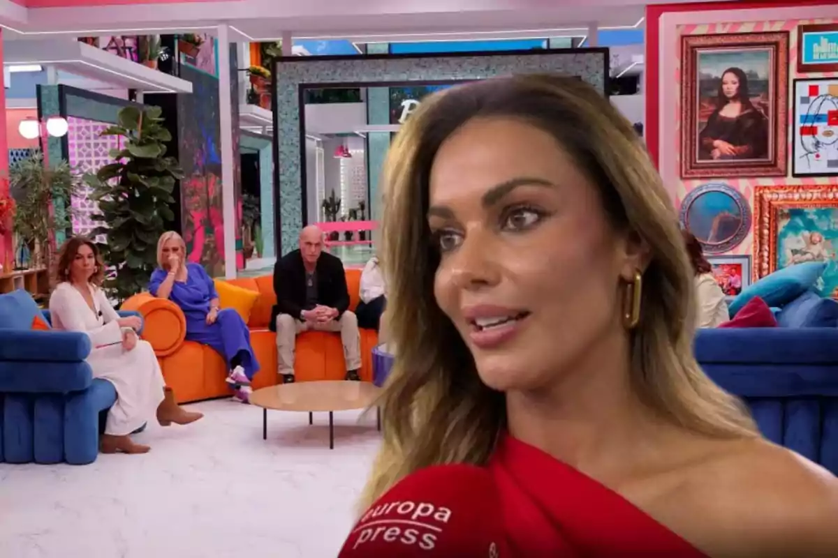 Una mujer con vestido rojo es entrevistada en primer plano mientras varias personas están sentadas en sofás de colores en un set de televisión decorado con cuadros y plantas