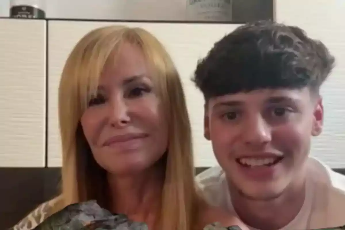 Una mujer rubia y un joven de cabello oscuro sonríen juntos frente a la cámara en un ambiente interior. Una mujer rubia y un joven de cabello oscuro sonríen juntos frente a la cámara en un ambiente interior.