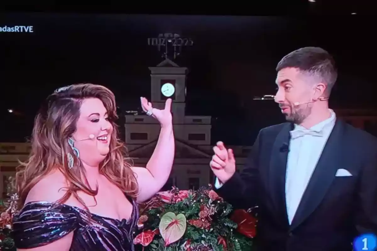Dos presentadores sonríen mientras están en un set decorado con flores y un reloj iluminado en el fondo. Dos presentadores sonríen mientras están en un set decorado con flores y un reloj iluminado en el fondo.