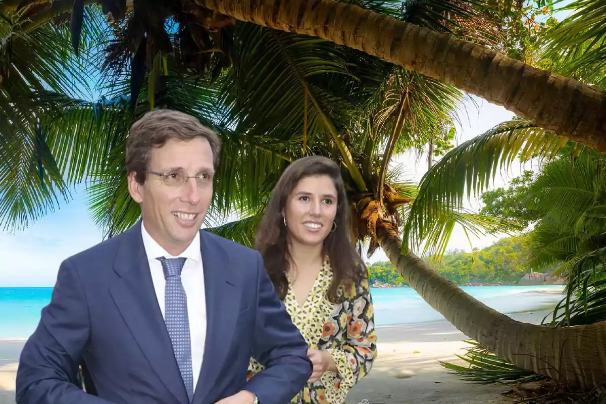 Una pareja sonriente, él con traje y corbata y ella con un vestido estampado, posando frente a una playa tropical con palmeras y mar azul.