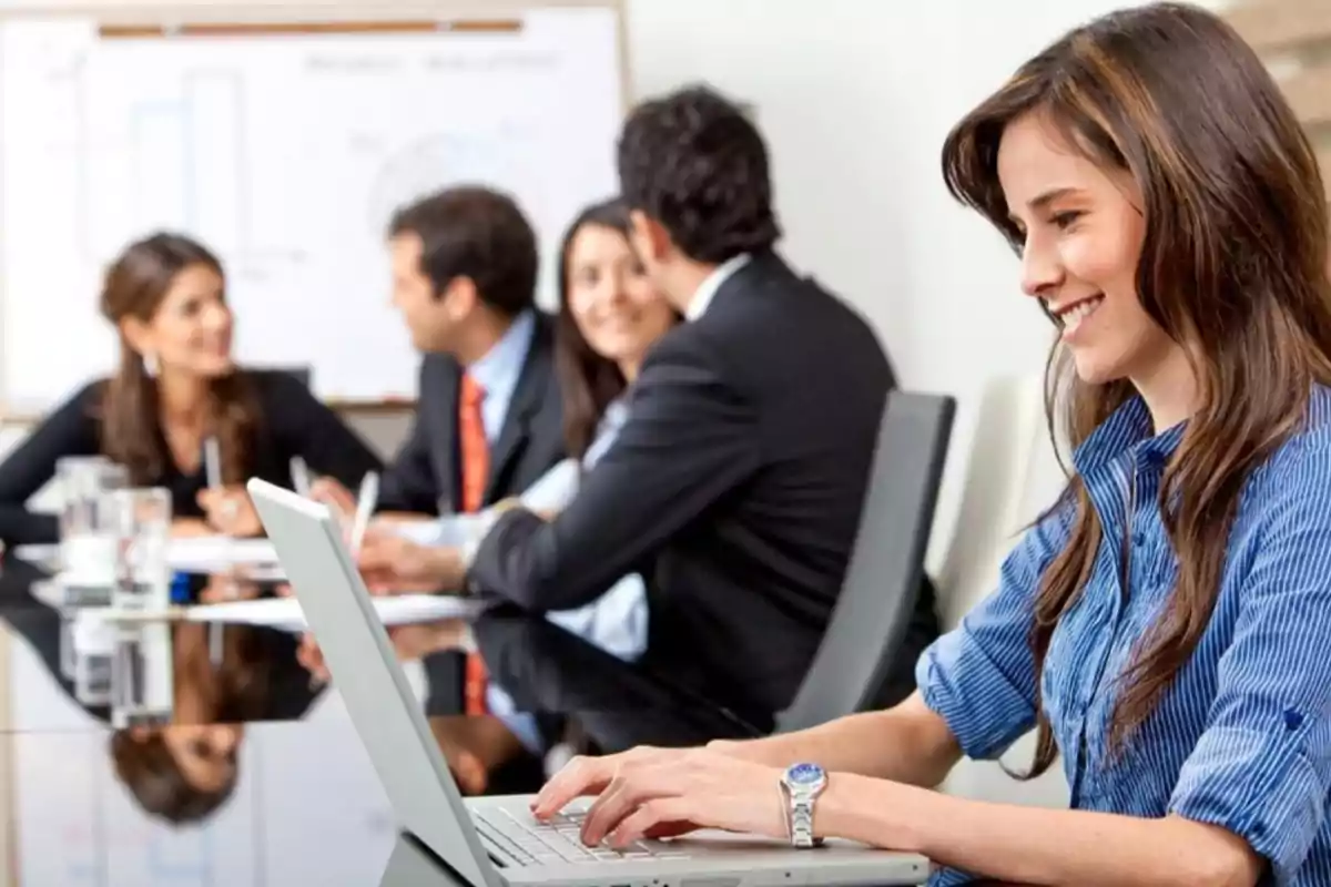 Una mujer sonriente trabaja en una computadora portátil mientras un grupo de personas se reúne en una sala de conferencias.