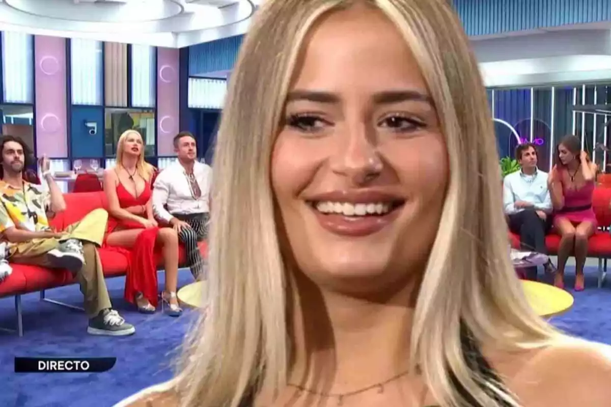 Una mujer rubia sonriente en primer plano con un grupo de personas sentadas en un sofá rojo en un plató de televisión.